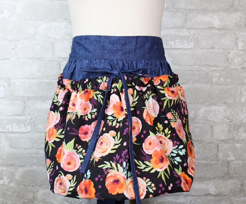 May include: A waist apron featuring a dark blue denim waistband with matching tie strings and a gathered skirt section. The skirt is made of black fabric printed with a vibrant floral pattern in shades of orange, pink, and light green foliage.