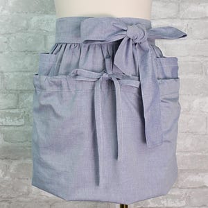 May include: A light blue waist apron featuring a gathered skirt design with a wide waistband and a large fabric tie at the center. The apron includes two side pockets and a secondary tie closure positioned lower on the front panel of the garment.