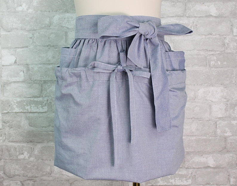 May include: A light blue waist apron featuring a gathered skirt design with a wide waistband and a large fabric tie at the center. The apron includes two side pockets and a secondary tie closure positioned lower on the front panel of the garment.