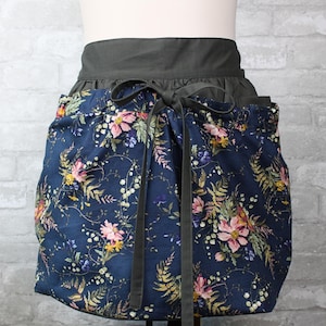 May include: A waist apron featuring a dark blue fabric base with a multi-colored floral and fern pattern in shades of pink, yellow, and green. The apron has a solid dark gray waistband and matching dark gray fabric ties that are tied in a bow.