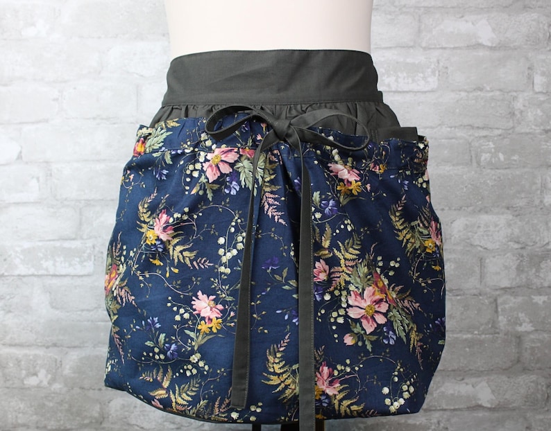 May include: A waist apron featuring a dark blue fabric base with a multi-colored floral and fern pattern in shades of pink, yellow, and green. The apron has a solid dark gray waistband and matching dark gray fabric ties that are tied in a bow.