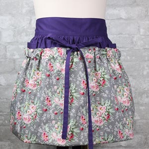 May include: A waist apron featuring a solid deep purple waistband with matching fabric ties. The lower section is made of a grey fabric printed with a floral pattern of pink roses and green leaves, gathered at the waist with a ruffled top edge.