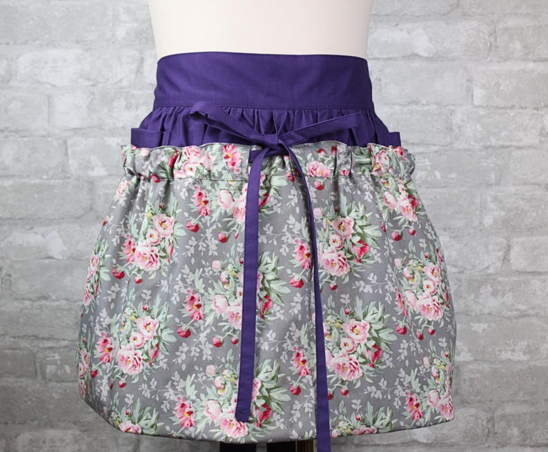 May include: A waist apron featuring a solid deep purple waistband with matching fabric ties. The lower section is made of a grey fabric printed with a floral pattern of pink roses and green leaves, gathered at the waist with a ruffled top edge.