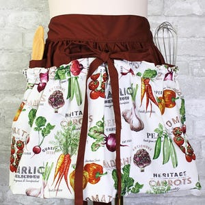 May include: A waist apron featuring a white fabric with a colorful vegetable print including carrots, tomatoes, garlic, and peas, paired with a solid dark brown waistband and tie. The apron includes two side pockets holding a wooden spoon and a whisk.