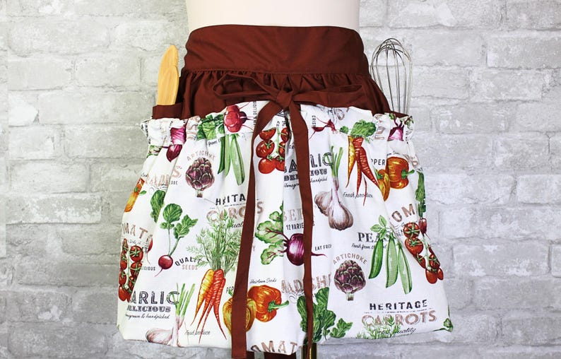 May include: A waist apron featuring a white fabric with a colorful vegetable print including carrots, tomatoes, garlic, and peas, paired with a solid dark brown waistband and tie. The apron includes two side pockets holding a wooden spoon and a whisk.