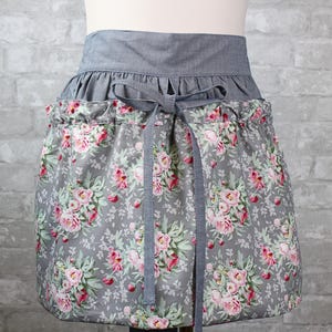 May include: A waist apron featuring a gray fabric waistband with a matching tie closure. The lower portion of the apron is made of a gray fabric printed with a floral pattern of pink roses and green leaves, gathered at the waist with a drawstring.