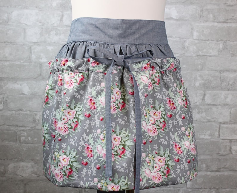 May include: A waist apron featuring a gray fabric waistband with a matching tie closure. The lower portion of the apron is made of a gray fabric printed with a floral pattern of pink roses and green leaves, gathered at the waist with a drawstring.