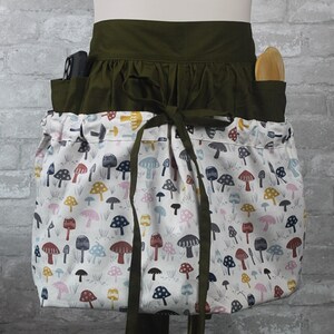 May include: A waist apron featuring a dark olive green waistband with matching ties and a large, gathered front pocket panel. The pocket fabric is white and printed with a repeating pattern of small mushrooms in shades of blue, pink, red, and yellow.