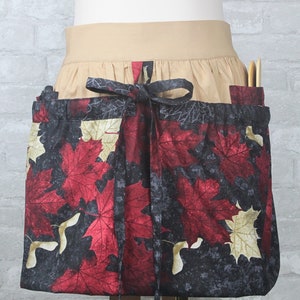May include: A waist apron featuring a black background with a pattern of red and cream-colored maple leaves and seed pods. The apron has a wide tan waistband with a gathered skirt section and a matching fabric tie closure at the center front.