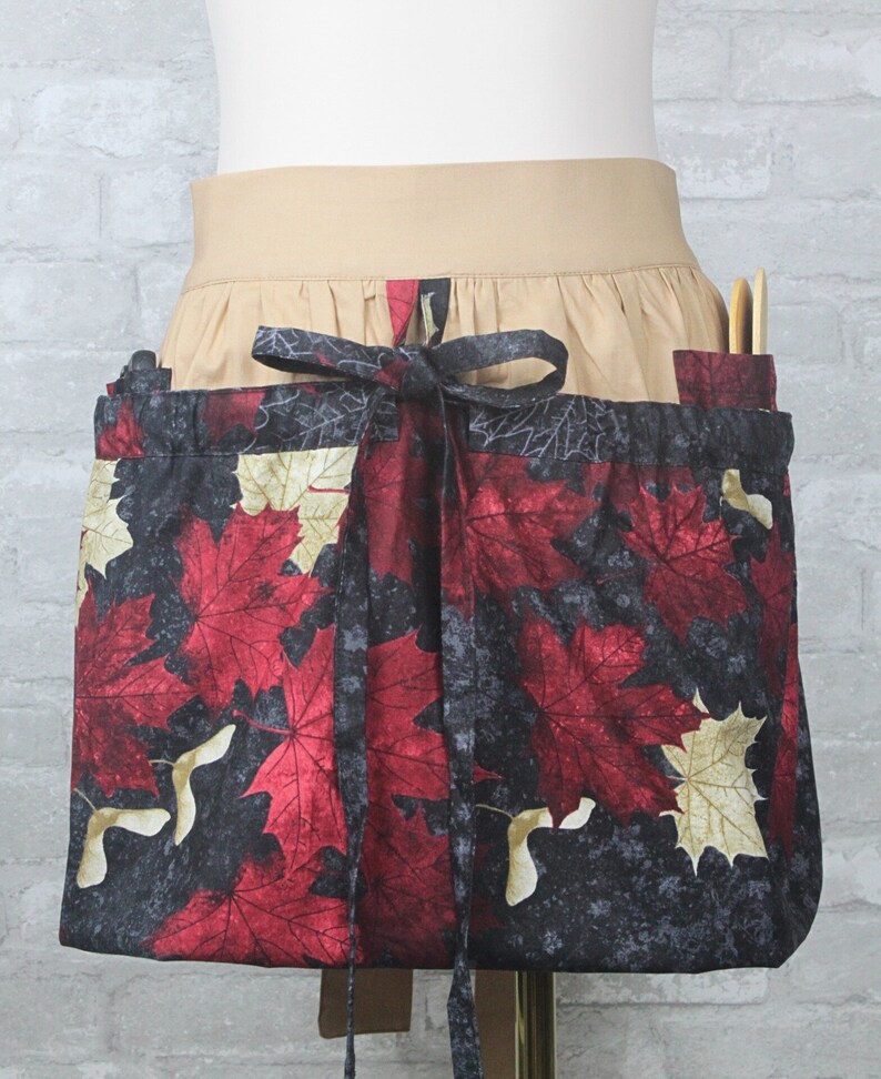 May include: A waist apron featuring a black background with a pattern of red and cream-colored maple leaves and seed pods. The apron has a wide tan waistband with a gathered skirt section and a matching fabric tie closure at the center front.