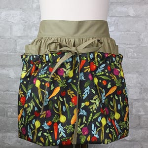 May include: A waist apron featuring a solid olive green waistband with a tie closure and a gathered skirt. The skirt is black and decorated with a colorful pattern of various vegetables including orange carrots, red tomatoes, and green leafy herbs.