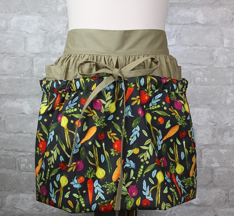 May include: A waist apron featuring a solid olive green waistband with a tie closure and a gathered skirt. The skirt is black and decorated with a colorful pattern of various vegetables including orange carrots, red tomatoes, and green leafy herbs.