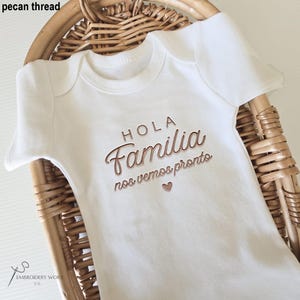 May include: White baby onesie with the words "HOLA Familia nos vemos pronto" embroidered in a copper-colored thread. The onesie is displayed in a woven basket. The text includes a heart symbol.