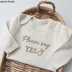 Puede incluir: Body de bebé color crema con mangas largas. El texto "Please say YES" está bordado en marrón, junto con un gráfico de anillo. El body se muestra en una cesta de mimbre.