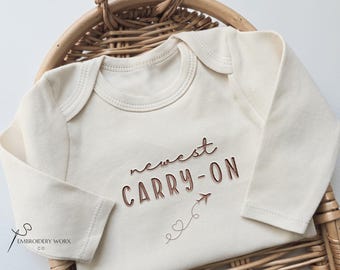May include: Cream-colored baby onesie with long sleeves, featuring the text "newest CARRY-ON" in brown script, along with a small airplane and heart design. The onesie is displayed in a woven basket.
