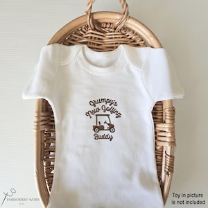 May include: A white baby onesie with the embroidered text "Grumpy's New Golfing Buddy" and a golf cart graphic in brown. The onesie is displayed in a woven basket.