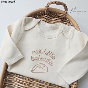 May include: Cream-colored baby onesie with long sleeves. The onesie features the embroidered text "our little baleada" with a taco illustration in beige thread. The onesie is displayed in a woven basket.