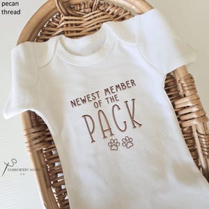 May include: White baby onesie with the text "NEWEST MEMBER OF THE PACK" in brown lettering, along with two paw prints. The onesie is displayed in a woven basket.
