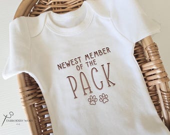 May include: White baby onesie with the text "NEWEST MEMBER OF THE PACK" in brown lettering, along with two paw prints. The onesie is displayed in a woven basket.