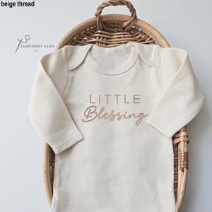 May include: Cream-colored baby onesie with long sleeves, featuring the embroidered text "LITTLE Blessing" in a soft, warm beige thread. The onesie is displayed in a woven basket, showcasing a simple, elegant design.