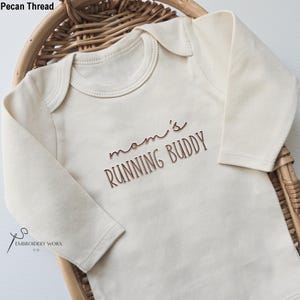 May include: Cream-colored baby onesie with long sleeves. The text "mom's RUNNING BUDDY" is embroidered in a brown thread across the front. The onesie is displayed in a woven basket.