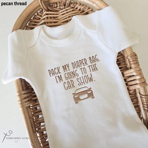 May include: White baby onesie with the embroidered text "PACK MY DIAPER BAG. I'M GOING TO THE CAR SHOW." and a car graphic. The text and graphic are in a pecan brown thread. The onesie is displayed in a wicker basket.