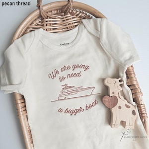 May include: Cream-colored baby onesie with the embroidered phrase "We are going to need a bigger boat" and a boat graphic in pecan thread. A wooden giraffe toy with a heart is next to the onesie. The onesie is in a woven basket.