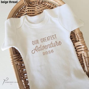 May include: A white baby onesie with short sleeves and the embroidered text "OUR GREATEST Adventure 2026" in beige thread. The onesie is displayed in a woven basket. The logo of "XS Embroidery Work co." is in the bottom left corner.