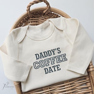 May include: Cream-colored baby onesie with long sleeves, featuring the text "DADDY'S COFFEE DATE" in a bold, outlined font. The onesie is displayed in a woven basket. The logo of "Embroidery Works Co." is visible in the lower left corner.