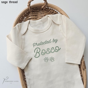 May include: Cream-colored baby onesie with long sleeves, featuring the text "Protected by Bosco" in green script, along with two paw prints. The onesie is displayed on a woven basket.