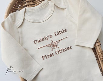 Peut inclure: Body de bébé crème à manches longues, avec l'inscription brodée "Daddy's Little First Officer" en fil marron. Le motif comprend un dessin d'avion. Le body est présenté dans un panier en osier.