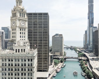May include: An aerial view of Chicago's cityscape with a river running through it. The river is lined with boats and bridges. The buildings are tall and modern, with a clock tower in the foreground.