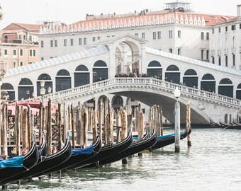 May include: A row of black gondolas with blue trim are docked in a canal in Venice, Italy. The iconic Rialto Bridge is visible in the background.