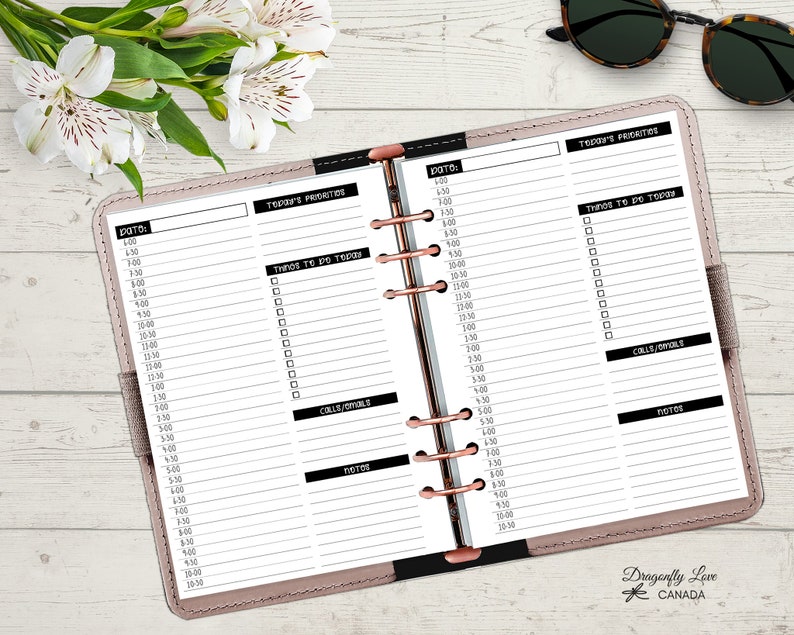 May include: Open planner with lined pages for daily scheduling. The pages are divided into sections for time slots, priorities, to-do lists, calls/emails, and notes. The planner is open on a light-colored surface with flowers and sunglasses.