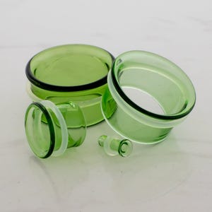 Op de afbeelding: Een verzameling groene glazen oorpluggen in verschillende maten. De pluggen hebben een afgeronde vorm met een heldere, lichtgroene tint. De set bevat een paar grotere pluggen, een kleinere plug en een enkele plug.