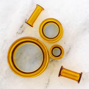 May include: A collection of amber-colored ear gauges in various sizes and shapes. The gauges include circular and cylindrical designs, with a glossy finish. The items are arranged on a white marble surface, showcasing their warm tones and smooth texture.