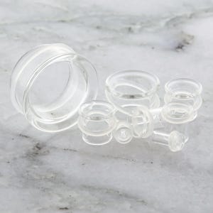 May include: Clear glass ear gauges of various sizes are displayed. The gauges have a tunnel design, with a rounded, cylindrical shape. The glass is transparent, allowing light to pass through. The gauges are arranged on a light-colored surface.