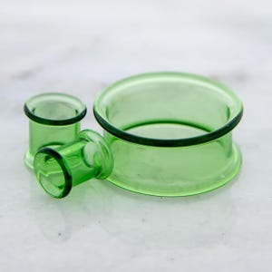 Peut inclure: Trois écarteurs d'oreille en verre vert translucide de différentes tailles sont présentés. Les écarteurs ont une forme cylindrique avec un bord évasé et une bordure vert foncé. Ils sont disposés sur une surface en marbre blanc, mettant en valeur leur texture lisse et polie.