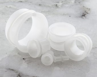 May include: A collection of white, translucent ear gauges and plugs in various sizes and styles. The assortment includes tunnel plugs, double-flared plugs, and a few smaller pieces, all made of a smooth, matte material.