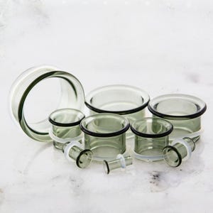 Puede incluir: Un conjunto de expansores de oreja de vidrio transparente, en varios tamaños. Los expansores tienen forma cilíndrica con un borde oscuro y un borde transparente ensanchado. Se exhiben sobre una superficie blanca.