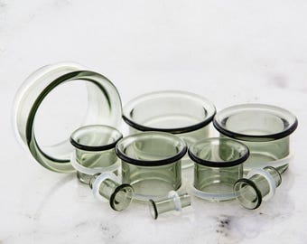 May include: A collection of clear glass ear gauges in various sizes. The gauges have a rounded, cylindrical shape with a dark rim and a clear, flared edge. The gauges are displayed on a white surface.
