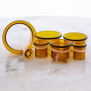 Puede incluir: Tapones y túneles para los oídos de vidrio color ámbar en varios tamaños. Los tapones tienen forma cilíndrica con un borde ensanchado, mientras que los túneles son circulares. Algunos tienen detalles en blanco y negro.