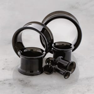 Puede incluir: Una colección de expansores de oreja negros de varios tamaños. Los expansores están hechos de un material brillante y tienen un diseño liso y redondeado. Los expansores se muestran sobre una superficie de mármol blanco.