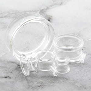 May include: A collection of clear glass ear gauges in various sizes. The gauges are cylindrical with flared edges, designed for stretched ear piercings. The transparent material allows light to pass through, highlighting their smooth, polished surfaces.