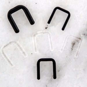 Puede incluir: Una colección de piercings nasales en forma de U, negros y transparentes. Los piercings están hechos de un material liso y están dispuestos sobre una superficie blanca veteada. La imagen muestra la variedad de colores y estilos disponibles.