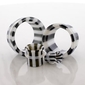 May include: Four pieces of body jewelry, including two large ear gauges, a small plug, and a small tunnel. The jewelry features a black and white striped pattern. The pieces are made of a translucent material and are arranged on a white surface.