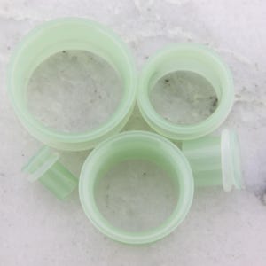 May include: A collection of light green ear gauges in various sizes. The gauges are made of a translucent material and have a smooth, rounded design. The ear gauges are displayed on a light-colored surface.