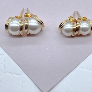 May include: A pair of gold wire wrapped pearl earrings. The pearls are white and the wire is gold. The earrings are on a light pink surface.