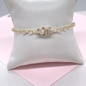May include: Gold-toned chain bracelet featuring a teddy bear charm. The charm is adorned with clear and pink crystals. The bracelet is displayed on a white pillow against a pink background, showcasing a delicate and feminine design.