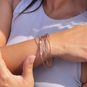 Könnte beinhalten: Nahaufnahme eines Handgelenks, das einen Stapel silberner und goldener Armreifen trägt. Die Armreifen sind dünn und zart und in einem Kreuzmuster angeordnet.